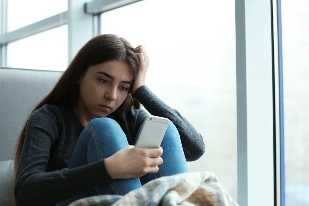 Bored teenage girl with smartphone sitting by window indoors