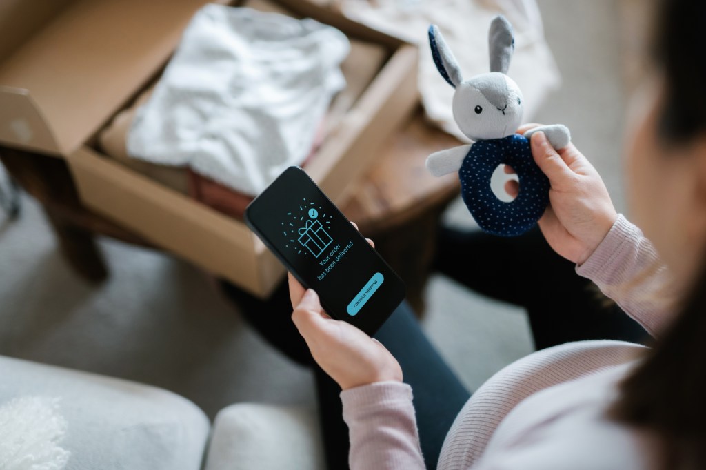 Over-the-shoulder view of a pregnant Asian woman on a sofa, shopping online on her smartphone and holding a bunny toy rattle, with a boxless delivery of baby items nearby