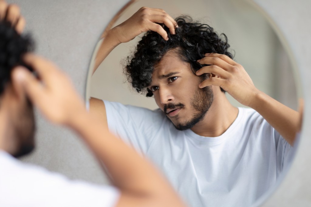 Worried Indian man examining his gray hair and scalp in the mirror at home