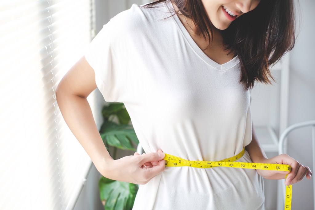 A woman smiles as she measures her waist. 