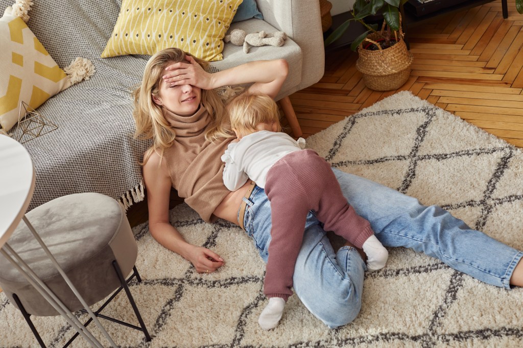 Caucasian blonde woman in jeans breastfeeding her baby while sitting on a rug in a bright, scandinavian style room